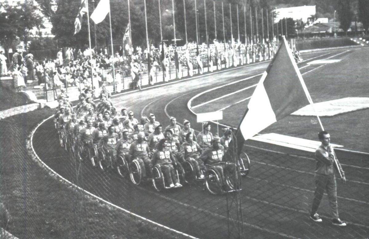 Imagem antiga, em preto e branco, que mostra uma pista de atletismo com várias pessoas sobre cadeira de rodas, em fileiras, seguindo um homem que caminha com a bandeira da Itália.