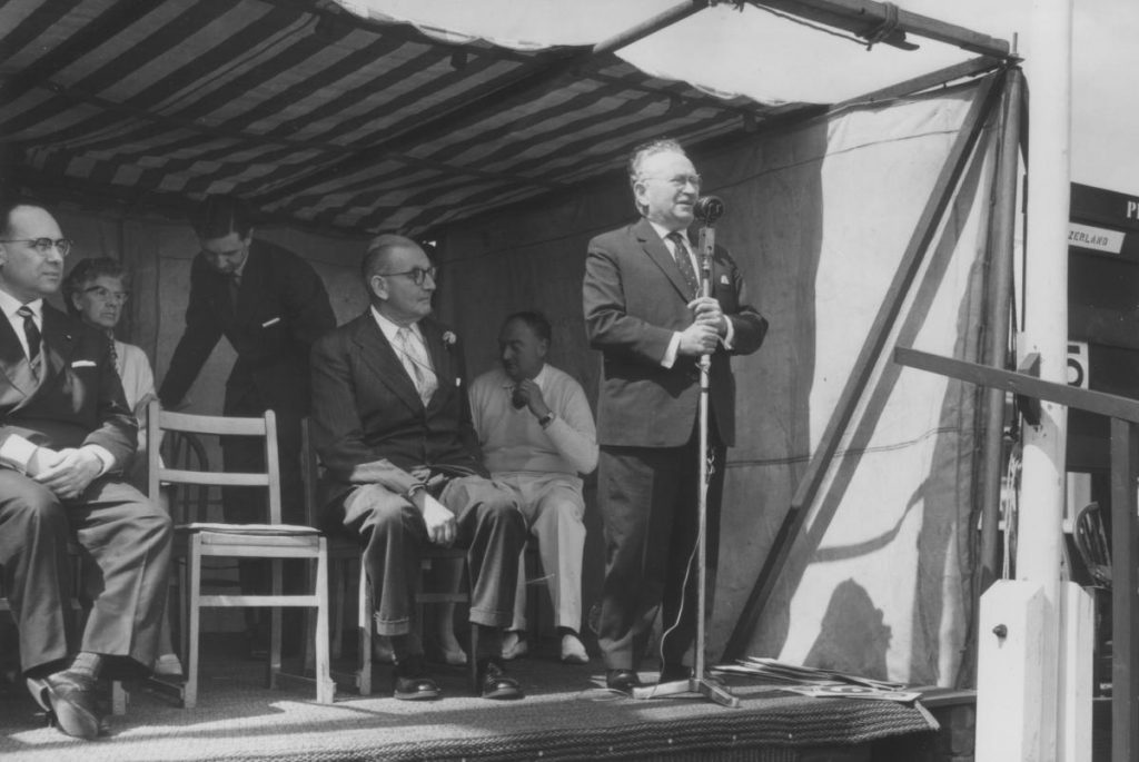 Fotografia antiga, em preto e branco, que mostra um homem falando ao microfone em pedestal. Ao lado dele há outro homem sentado.