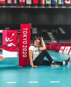 Luiza está sentada em quadra de volei, sorrindo para a cânera e encostada em uma placa escrita Tokyo 2020