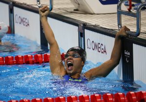 Cecilia é uma garota negra, ela está dentro de uma piscina e levanta um dos braços em comemoração, gritando feliz. Ela usa a touca e o óculos de natação.