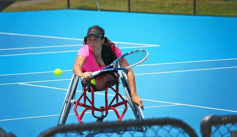Foto mostra uma jogadora de tenis sobre cadeira de rodas, em quadra, preparando a raquete para golpear uma bola de tenis