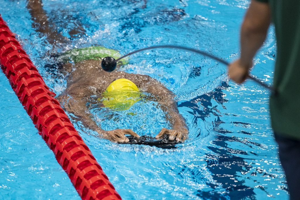 Nadador deficiente visual dentro de uma piscina frente a natação, com uma touca amarela, sendo guiado por seu ajudante com um instrumento chamado de "tapper".
