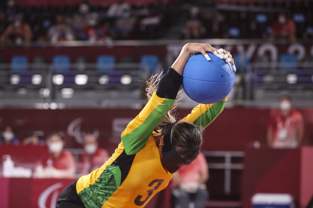 Foto de uma atleta durante o jogo de Golbol, ela usa a venda preta e agarra uma bola azuul, com os braços levantados.