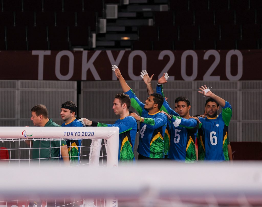 Seleção brasileira de GoalBall, com 6 atletas homens, aparece ao fundo da imagem, em uma fila indiana, cada um segurando no ombro do outro para caminharem com segurança. Os 3 últimos acenam com uma das mãos para o alto. 