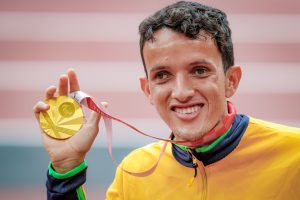 Imagem de perfil do atleta Yeltsin, um homem branco, sorridente, segurando a medalha de ouro com sua mão direita.