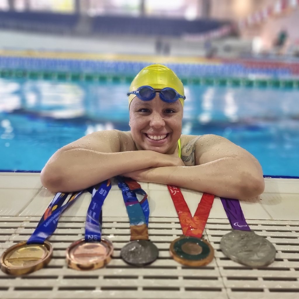 foto perfil de Edênia posando sobre a borda de uma piscina. Em frente a ela, estão 5 medalhas de competições diferentes: uma de ouro, duas de bronze e duas de prata. 