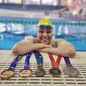 foto perfil de Edênia posando sobre a borda de uma piscina. Em frente a ela, estão 5 medalhas de competições diferentes: uma de ouro, duas de bronze e duas de prata.