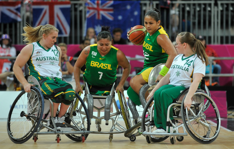 Quatro jogadoras de basquete em cadeira de rodas estão em quadra disputando uma bola que está na mãos de uma das atletas. Duas delas vestam o uniforme da seleção brasileira, incluindo a que está com a bola na mão e as outras duas são da seleção australiana.