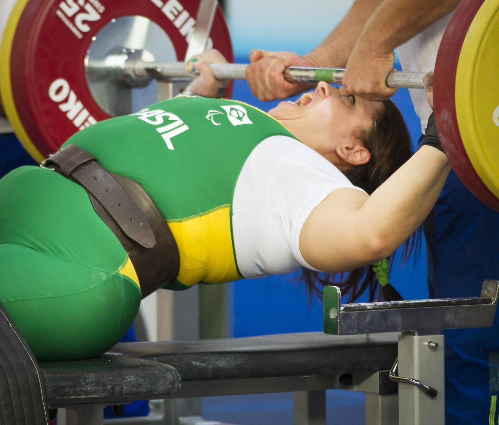 Atleta deitada em um banco carrega um peso e grita. Ela veste o uniforme verde da seleção brasileira.