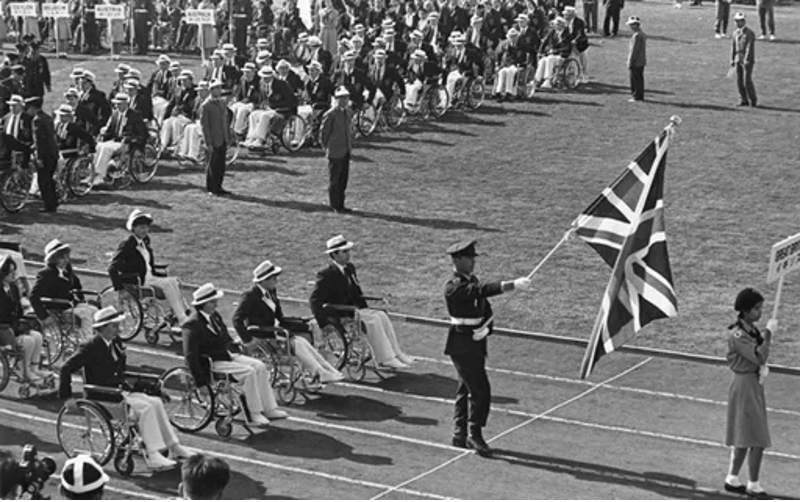 Foto em preto e branco de um desfile com várias pessoas agrupadas sobre cadeiras de rodas em uma pista de atletismo e um chapeu coco n