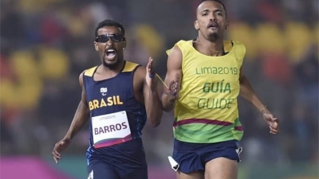 2 atletas negros brasileiros, um deles uma óculos e está ao lado de seu atleta-guia, que o auxilia na corrida referente a modalidade do atletismo. Ambos ligados um ao outro no punho com uma fita.