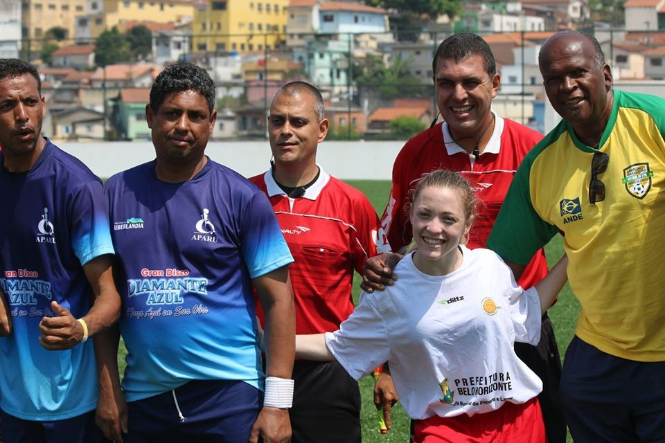 Foto que mostra 5 homens uniformizados junto à atleta Mariana Damásio, a mais baixa deles. Ela aparece sorrindo e junto com eles fazem pose para foto.