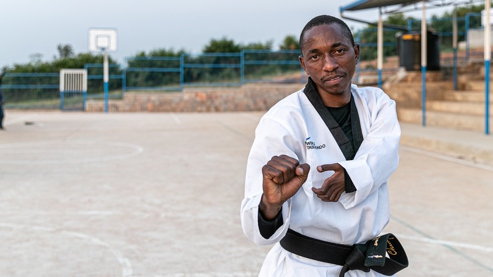 Parfai é um homem negro. Ele está em um local aberto e ensolarado, vestindo um dobok, roupão branco típico da luta de taekwondo.