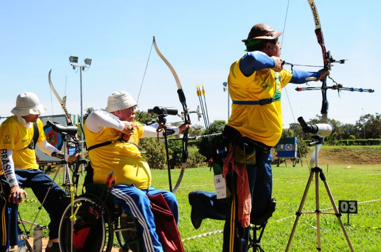 Foto mostra três atletas do tiro com arco, um deles sobre cadeira de rodas. Dois deles preparam o arco para atirar.