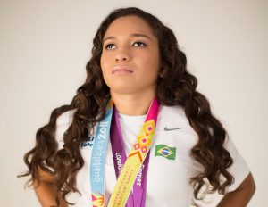 Jhulia, com a cabeça um pouco levantada virada para a diagonal, veste camisa branco e tem três medalhas no pescoço.