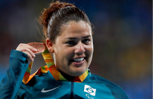 Atleta Marivana Oliveira da Nóbrega, mulher branca, cabelos castanhos, vestida com um casaco azul marinho, sorri enquanto segura com a mão direita a medalha de bronze. O fundo da imagem está desfocado. Foto: CPB