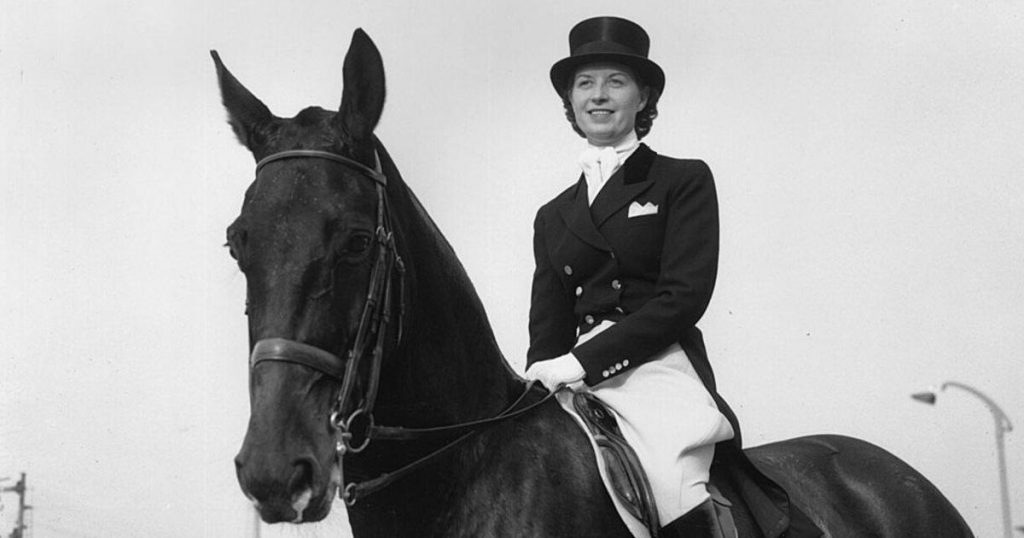 Lis está sobre um cavalo, em uma foto em preto e branco. Ela sorri e usa cartola e paletó.