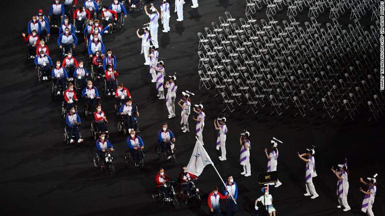 Foto da cerimônia de abertura dos jogos paralímpicos de Tóquio, quando a delegação russa desfila com diversos participantes em cadeiras de rodas. Na lateral à direita aparece uma série de cadeiras vazias.