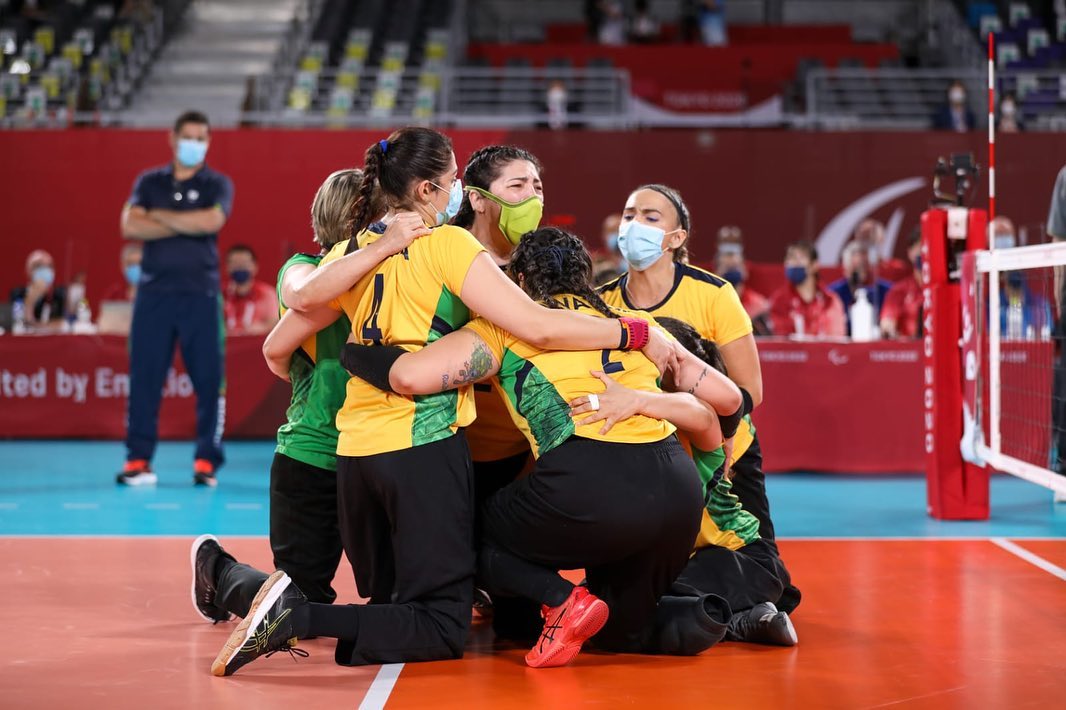 Foto da seleção feminina de volei sentado, sentadas na quadra de Tóquio, abraçadas em roda, todas usando máscaras.