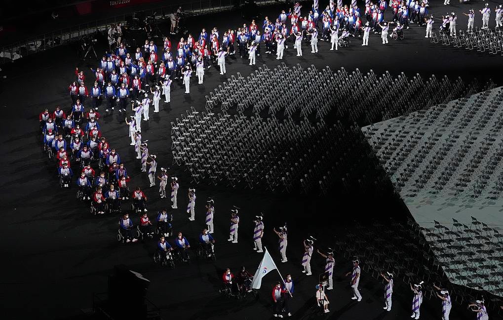 Foto da cerimônia de abertura dos jogos paralímpicos de Tóquio, quando a delegação russa desfila com diversos participantes em cadeiras de rodas. Na lateral à direita aparece uma série de cadeiras vazias.