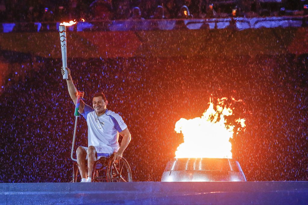 Descrição do Post: #PraTodoMundoVer: Clodoaldo (à esquerda) bosa sobre o palco branco, ao lado da pira paraolímpica do Rio 2016. Ele usa roupas brancas e está sobre a sua cadeira de rodas. Na imagem está de noite e chove, e as chamas fortes iluminam o cenário.