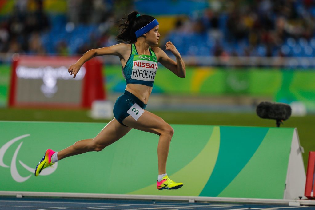 Verônica está em movimento, de lado, correndo em uma pista de atletismo. Ela veste camiseta e shorts da delegação. 