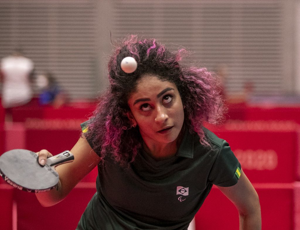 Foto de Milena durante a partida de tenis de mesa. Ela uma garota negra, com os cabelos crespos pintados de rosa.