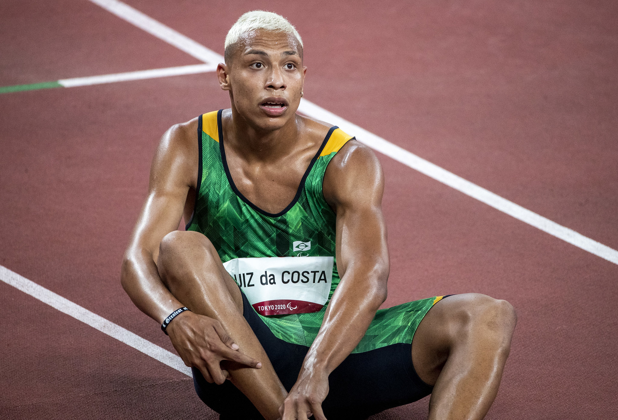 Christian é um rapaz negro, loiro e está sentado na pista de atletismo de Tóquio após uma corrida.