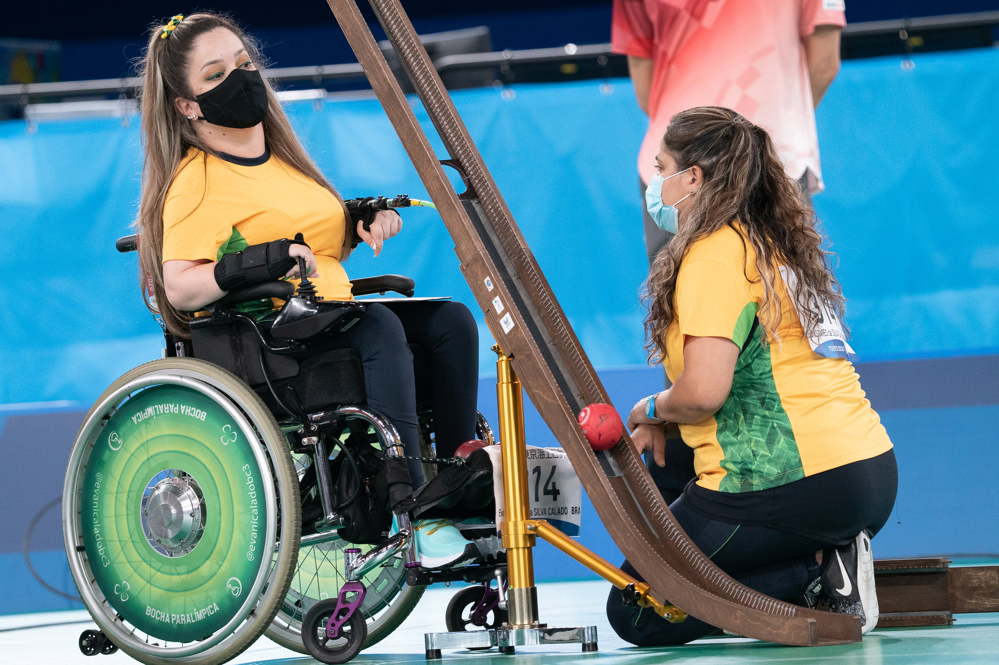 Foto que mostra Evani, na cadeira de rodas, com a camisa da seleção brasileira, de máscara e diante de uma calha, instrumento usado para arremar a bola da bocha. Ela está na frente de sua guia, que está ajoealhada.