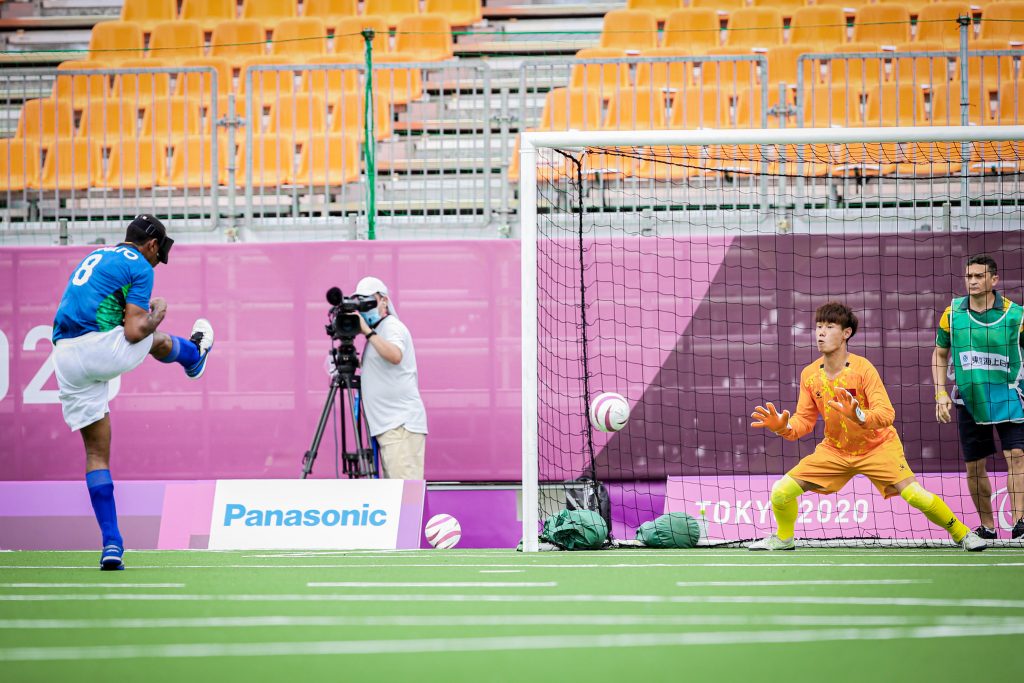 Foto de uma partida de futebol de 5 no momento em que o brasileiro chuta a bola ao gol contra o Japão. Atrás do gol está o guia ou chamador. 