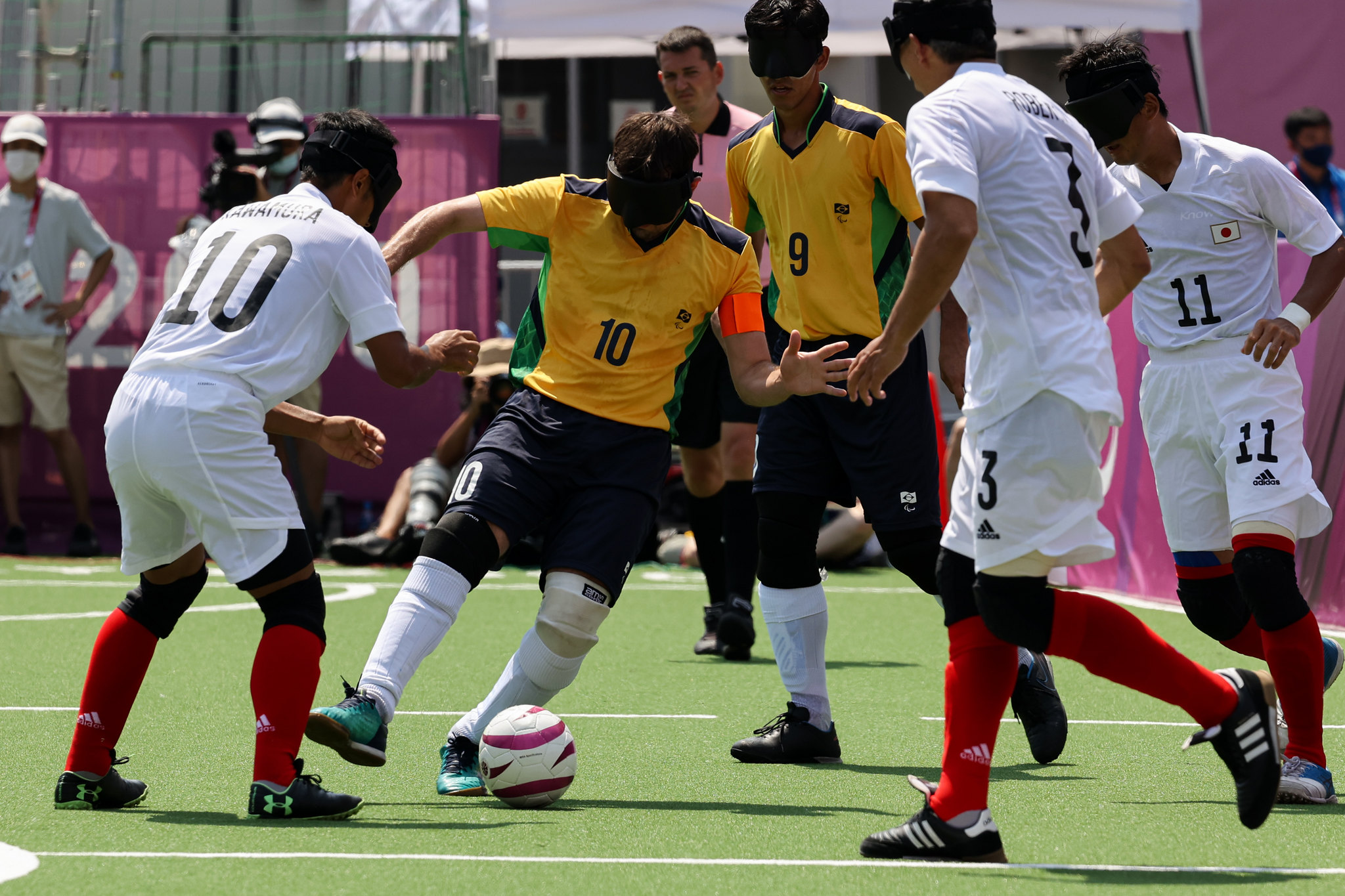 Foto de uma partida de futebol contra o Japão em Tóquio.Há dois brasileiros, um deles com domínio da bola, e outros dois japoneses tentando alcançá-la. Todos com venda nos olhos.