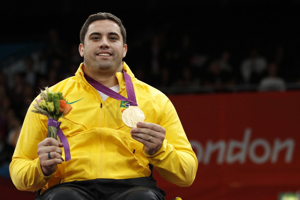 Homem branco de cabelo castanho preto, usando um agasalho todo amarelo representando o Brasil, em uma mão segurando a medalha de ouro conquistada, e um ramo flores na outra simbolizando a honra dos jogos paralímpicos.