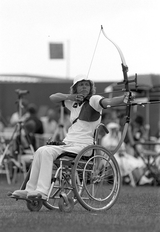 Foto em preto e branco mostra Neroli sobre cadeira de rodas apontando seu arco