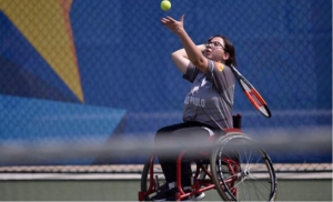 Foto de Fernanda, mulher branca, cabelos curtos, castanhos, olhos da mesma cor. Ela esta sentada em sua cadeira, que possui detalhes vermelhos e pretos, jogando tênis em uma quadra, ela está neste momento jogando a bola para o saque.