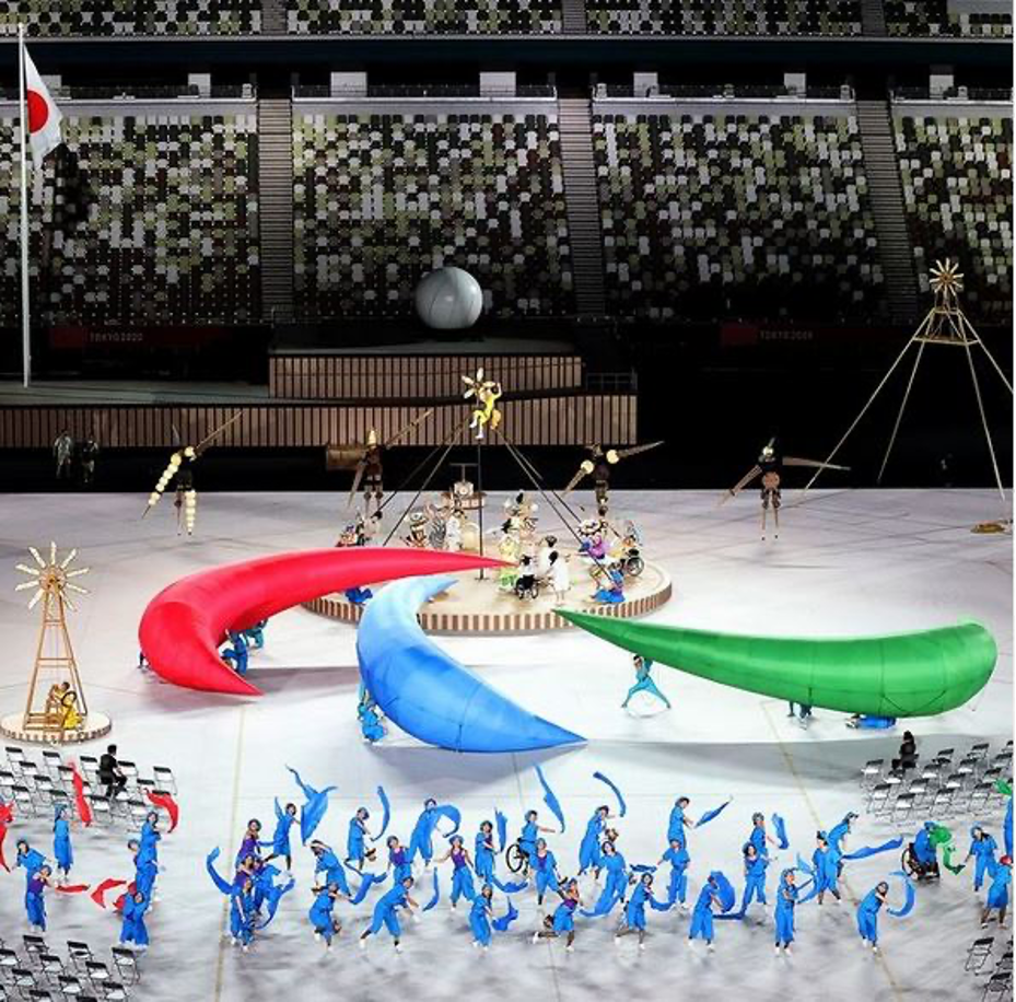 foto da cerimônia de abertura da Paralimpíada onde está mostrando uma performance artística feita por várias pessoas. No centro do palco, os artistas carregam três arcos de espuma em vermelho, verde e azul que juntos formam o “Agitos”, símbolo paralimpíco.