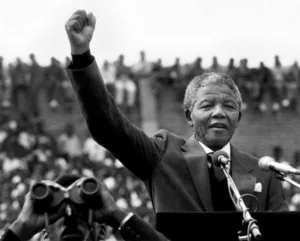 nelson mandela com o punho cerrado e o braço levantado apontando para o céu fazendo um discurso para uma multidão