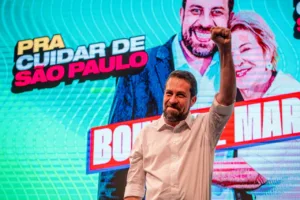 Guilherme Boulos está com camisa branca, em frente a telão de led com imagem dele e de Marta Suplicy ao fundo, com uma mão para cima e punho em riste.