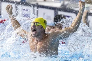 Na imagem, o nadador paralímpico Douglas Matera está dentro da piscina, com os braços erguidos e punhos cerrados, expressando intensa comemoração. Ele usa uma touca amarela e óculos de natação com lentes amarelas. Sua expressão facial é de euforia, com a boca aberta em um grito de celebração. A água ao seu redor está agitada, criando respingos altos, evidenciando o movimento explosivo de sua vitória. O cenário ao fundo mostra uma competição, com outros nadadores parcialmente visíveis, mas o foco é em Douglas, celebrando seu desempenho.