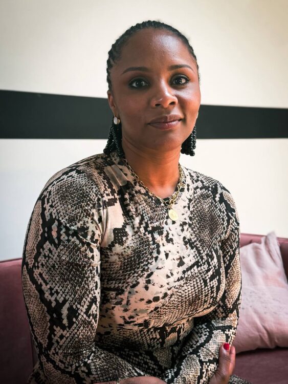 Gessica Généus, diretora e protagonista de Marie Madeleine, sentada em um sofá e olhando diretamente para a câmera. Ela é uma mulher negra, usa cabelo preso com tranças, brincos pequenos, colar dourado e uma blusa de manga longa com estampa preta e branca. A luz entra pela lateral e ilumina seu rosto em um retrato feito em ambiente interno.