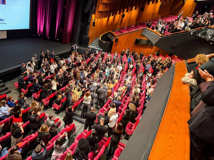 Sala de cinema do Festival de Cannes vista de cima, com poltronas vermelhas, paredes de madeira e a plateia em pé aplaudindo após a exibição. No centro, a equipe do filme Marie Madeleine recebe a ovação cercada pelo público.